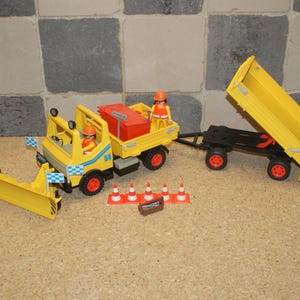 Op de afbeelding: Een gele speelgoedvrachtwagen met een sneeuwploeg, aanhanger en twee speelfiguren. De vrachtwagen heeft een gele cabine met blauw-witte geblokte accenten. De aanhanger is zwart met een gele laadbak. Inclusief oranje verkeerskegels.