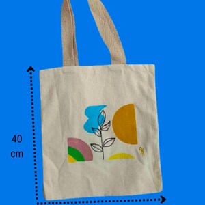 May include: A white canvas tote bag with a colourful abstract design featuring a blue flower, yellow and orange shapes, and green and pink accents. The bag measures 37 cm wide and 40 cm tall.