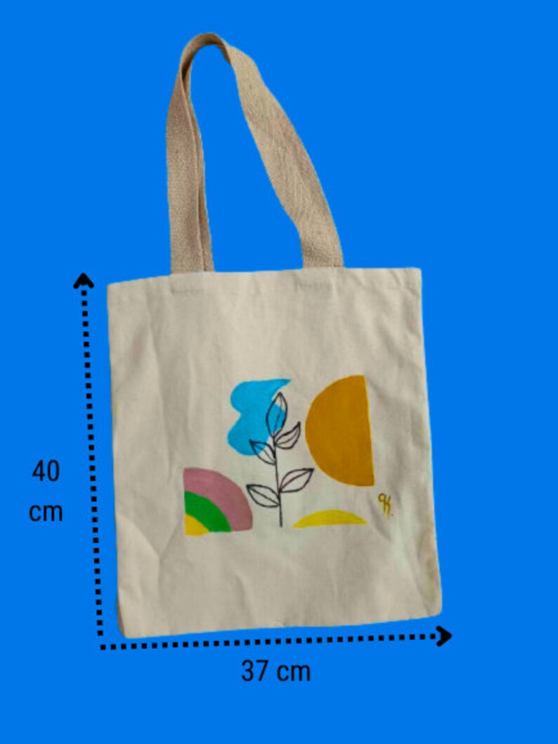May include: A white canvas tote bag with a colourful abstract design featuring a blue flower, yellow and orange shapes, and green and pink accents. The bag measures 37 cm wide and 40 cm tall.