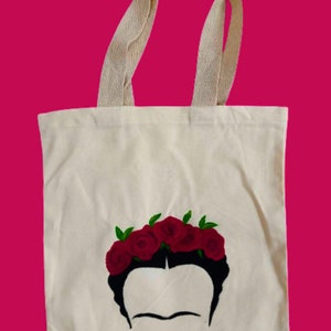May include: A white canvas tote bag featuring a black and white illustration of Frida Kahlo wearing a crown of red roses. The tote bag is made from natural cotton fabric and has two long cotton straps.