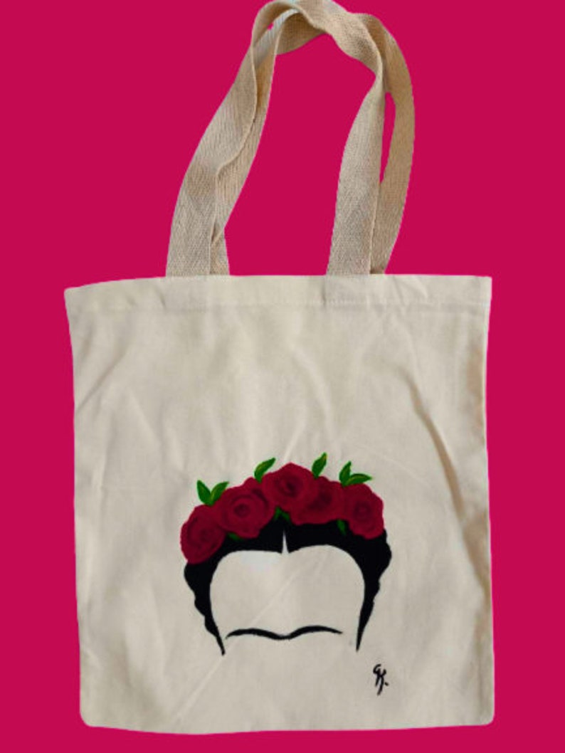 May include: A white canvas tote bag featuring a black and white illustration of Frida Kahlo wearing a crown of red roses. The tote bag is made from natural cotton fabric and has two long cotton straps.