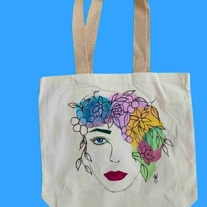 May include: A white canvas tote bag featuring a colourful floral design with a woman's face. The flowers are painted in shades of pink, blue, yellow, and green.