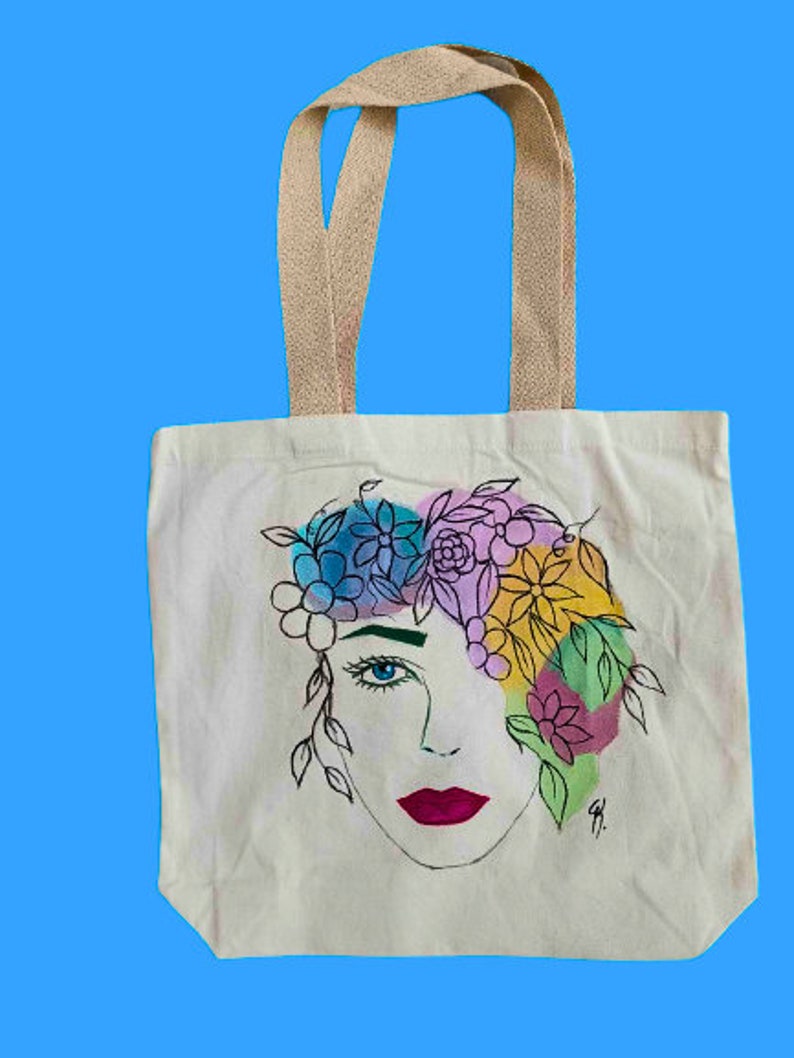 May include: A white canvas tote bag featuring a colourful floral design with a woman's face. The flowers are painted in shades of pink, blue, yellow, and green.