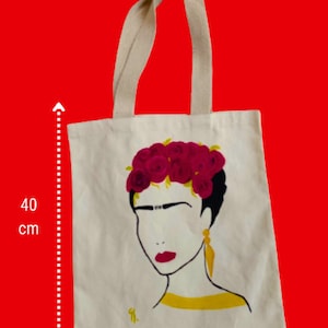 May include: A white canvas tote bag featuring a black and yellow illustration of Frida Kahlo with a red flower crown. The bag measures 40 cm tall and 37 cm wide.
