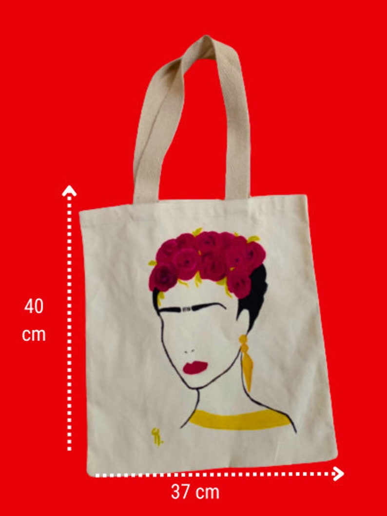 May include: A white canvas tote bag featuring a black and yellow illustration of Frida Kahlo with a red flower crown. The bag measures 40 cm tall and 37 cm wide.