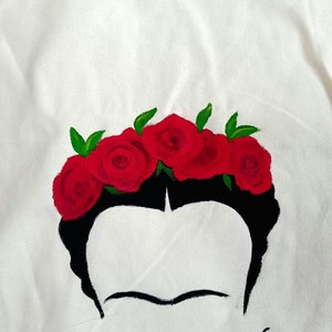 May include: A white canvas tote bag with a black and red painted design. The design features a stylised portrait of Frida Kahlo with a crown of red roses.