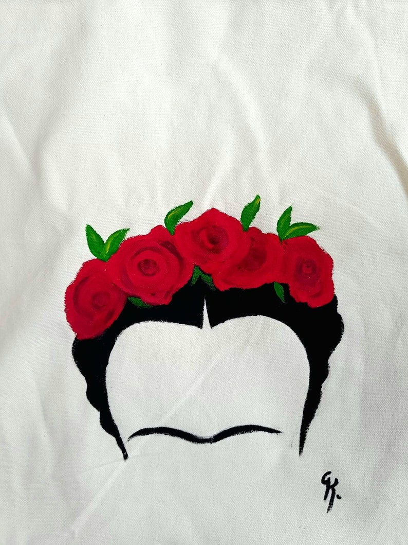 May include: A white canvas tote bag with a black and red painted design. The design features a stylised portrait of Frida Kahlo with a crown of red roses.