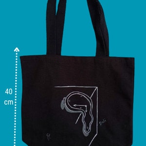 May include: Black canvas tote bag featuring a white illustration of a melting clock and the word "Dali" on the front. The bag measures 37 cm wide and 40 cm tall.
