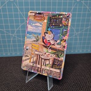 May include: A holographic Pokémon trading card featuring Alolan Meowth relaxing at a beachside cafe. The card has a colorful illustration with the text "Alolan Meowth" and other game-related details. The card is displayed on a clear stand.