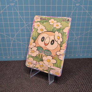 May include: A Rowlet Pokémon trading card, featuring a cartoon owl character with large eyes and a floral design. The card is displayed on a clear plastic stand against a blue grid background. The card has a HP of 60.