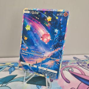 Könnte beinhalten: Holografische Minior Pokémon-Sammelkarte. Die Karte zeigt einen rosa und weißen Minior-Charakter mit sternenklarem Hintergrund. Die Karte lautet "Star Drop" und "HP 80".