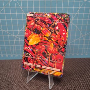 May include: A Buzzwole EX Pokemon trading card. The card features a red and black design with a detailed illustration of the Buzzwole character. The card is displayed on a clear plastic stand.