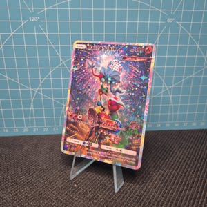 May include: A Blacephalon Pokémon trading card, displayed on a clear acrylic stand. The card features a colorful illustration of a creature with fireworks and carnival imagery. The card has a HP of 100 and a damage of +20.