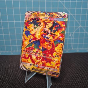 Könnte beinhalten: Eine Mega Charizard Y EX Pokémon-Karte mit einer feurigen Illustration in Orange, Rot und Blau. Die Karte zeigt den Text "Crimson Dive" und "HP 220". Sie wird auf einem transparenten Kunststoffständer präsentiert.