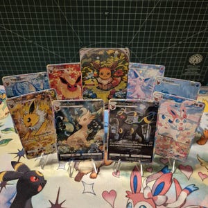 Peut inclure: Collection de cartes à collectionner Pokémon, avec Eevee, Jolteon et Umbreon, présentées sur des présentoirs transparents. Les cartes présentent des illustrations colorées et des détails de personnages. Les cartes ont différentes valeurs de PV.