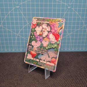 Può includere: Una carta collezionabile di Pokémon con il personaggio Maushold. La carta presenta un'illustrazione colorata di creature bianche simili a topi in un ambiente forestale. La carta è appoggiata su un supporto di plastica trasparente.