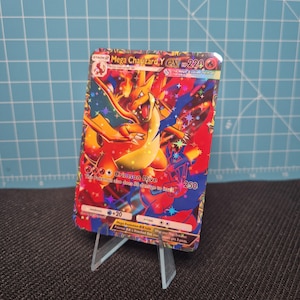 May include: A Mega Charizard Y EX Pokémon trading card. The card features a vibrant illustration of Charizard in shades of orange, red, and blue. The card displays the text "Crimson Dive" and "250". The card is displayed on a clear plastic stand.