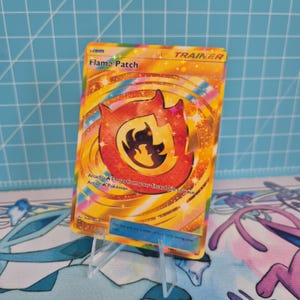 Könnte beinhalten: Eine goldene Trainerkarte mit einem feurigen Design. Die Karte zeigt eine große orangefarbene Flamme mit schwarzem Innenraum, und der Text "Flame Patch" ist sichtbar. Die Karte wird in einem transparenten Kunststoffständer gehalten.