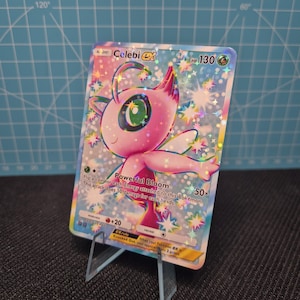 May include: A holographic Celebi ex Pokémon trading card. The card features a pink and green creature with the text "Powerful Bloom". The card is displayed on a clear acrylic stand.