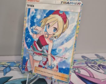 Könnte beinhalten: Eine holografische Sammelkarte mit der Figur Irida. Die Karte hat eine farbenfrohe Illustration einer jungen Frau mit blonden Haaren und einem roten Stirnband. Der Text auf der Karte enthält "Supporter", "Irida" und Spielanweisungen.
