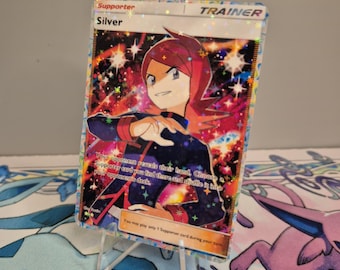 Puede incluir: Una tarjeta de Entrenador holográfica con el personaje Silver. La tarjeta tiene un fondo colorido con un diseño de galaxia. El texto de la tarjeta dice "Supporter" y "Trainer". La tarjeta se muestra en un soporte transparente.