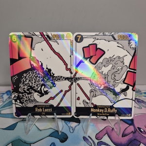 Peut inclure: Deux cartes à collectionner holographiques représentant des personnages de l'anime One Piece. Les cartes présentent Rob Lucci et Monkey D. Luffy. Chaque carte affiche un numéro, une puissance de 9000 et le nom du personnage. Elles sont présentées sur un support transparent.