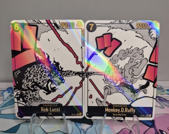 Könnte beinhalten: Zwei holografische Sammelkarten mit Charakteren aus dem Anime One Piece. Die Karten zeigen Rob Lucci und Monkey D. Luffy. Jede Karte hat eine Nummer, eine Stärke von 9000 und den Namen des Charakters. Sie werden auf einem transparenten Ständer präsentiert.