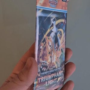 K&ouml;nnte beinhalten: Ein Pok&eacute;mon TCG Triumphant Light Themen-Boosterpack. Das Pack zeigt eine farbenfrohe Illustration einer wei&szlig;en und goldenen Kreatur mit blauem Hintergrund. Das Pack wird in einer Hand gehalten.