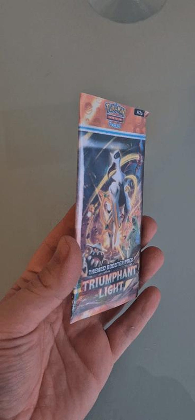 K&ouml;nnte beinhalten: Ein Pok&eacute;mon TCG Triumphant Light Themen-Boosterpack. Das Pack zeigt eine farbenfrohe Illustration einer wei&szlig;en und goldenen Kreatur mit blauem Hintergrund. Das Pack wird in einer Hand gehalten.
