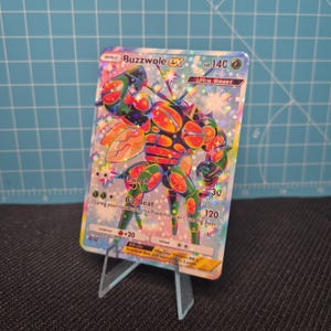 May include: A holographic Buzzwole EX Pokémon trading card, displayed on a clear acrylic stand. The card features a colorful illustration of the Ultra Beast character with the text "Buzzwole EX", "Punch", and "Big Beat".