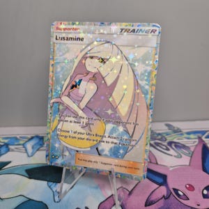 May include: A holographic Trainer card featuring the character Lusamine. The card has a silver border and a colorful design with stars and sparkles. The text on the card includes "Supporter", "Lusamine", and game instructions.