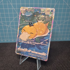 Puede incluir: Una carta coleccionable Pokémon holográfica de Psyduck. La carta presenta un personaje Psyduck amarillo con un pico blanco y un cuerpo verde, sobre un fondo de agua azul y verde. La tarjeta tiene el texto "Psyduck" y "Headache".