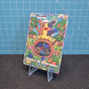 May include: A holographic Pokémon trading card featuring Morpeko, a yellow and black creature, with the text "Full Belly Bolt." The card is displayed on a clear plastic stand against a blue grid background. The card has a colorful, illustrated background.
