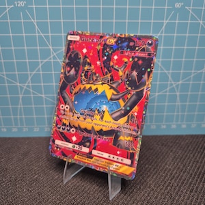 May include: A Guzzlord EX Pokémon trading card, featuring a red, black, and yellow monster illustration. The card displays text including "Grindcore" and "Tyrannical Hole", and is propped up on a clear plastic stand.