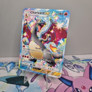 Puede incluir: Una carta coleccionable Pokémon Charizard EX. La carta presenta una ilustración estilizada de Charizard con cuerpo negro, vientre naranja y alas rojas. La carta tiene un fondo azul y rosa con el texto "Stoke" y "Steam Artillery".