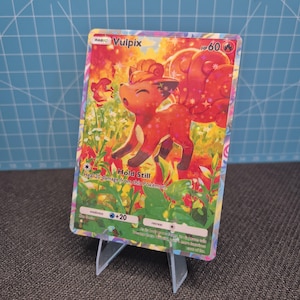 Peut inclure: Une carte à collectionner Vulpix avec une finition holographique représentant une créature ressemblant à un renard rouge dans un champ de fleurs jaunes et vertes. La carte affiche le nom Vulpix, 60 PV, un symbole feu et l'attaque Hold Still.