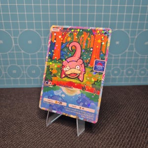 May include: A holographic Slowpoke Pokémon trading card. The card features a pink Slowpoke character in a forest scene with the word "Headbutt" and other game details. The card is displayed on a clear plastic stand.