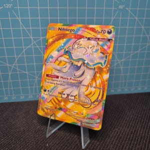 May include: A holographic Nihilego Pokémon trading card. The card features a colorful design with a central illustration of the Ultra Beast Nihilego. The card has text describing its abilities and stats, including "More Poison" and "New Wave".