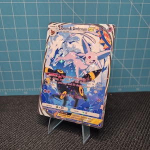 Puede incluir: Una carta coleccionable de Pokémon con Espeon & Umbreon EX. La carta tiene un diseño holográfico con un Espeon rosa y un Umbreon negro. La carta se muestra en un soporte de plástico transparente.