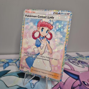 May include: A holographic Pokémon Center Lady card. The card features an illustration of a woman in a nurse's uniform with pink hair and blue eyes. The card has text that reads "Pokémon Center Lady" and "Trainer".