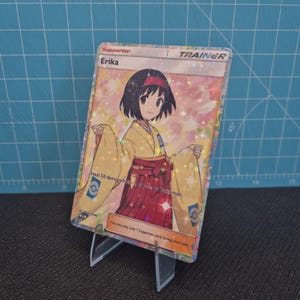 May include: A holographic Erika Trainer card from the Pokémon Trading Card Game. The card features an illustration of a young woman in a yellow kimono with red accents. The card is displayed on a clear plastic stand.