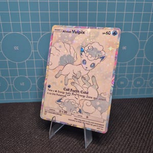 Könnte beinhalten: Eine holografische Alolan Vulpix Pokémon-Karte, präsentiert auf einem transparenten Ständer. Die Karte zeigt eine hellblaue und weiße Illustration des Pokémon mit dem Text "Call Forth Cold" und anderen Spieldetails. Die Karte befindet sich auf einem blauen Gitterhintergrund.