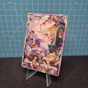 May include: A holographic Silvally Pokémon trading card, featuring a white and purple creature with a green and orange background. The card displays the text "Brave Buddies" and "HP 110". The card is displayed on a clear plastic stand.