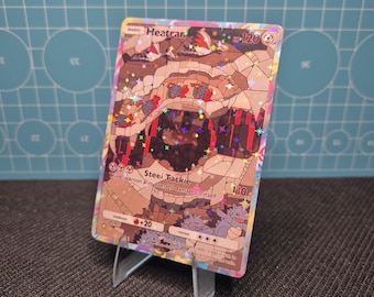 Puede incluir: Una carta Pokémon Heatran holográfica, exhibida en un soporte transparente. La carta presenta una ilustración detallada con tonos marrones, rojos y morados. El texto "Heatran" y "Steei Tackie" es visible.