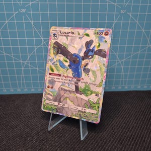 May include: A Pokémon trading card featuring Lucario, displayed on a clear acrylic stand. The card has a holographic design with a blue and black Lucario character. Text on the card includes "Lucario" and "HP 100". The background has a grid pattern.