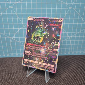 May include: A holographic Pokémon trading card featuring Celesteela, an Ultra Beast. The card has a green and yellow color scheme with a star-filled background. The card is displayed on a clear plastic stand.