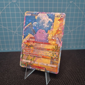 Könnte beinhalten: Holografische Ditto Pokémon-Sammelkarte. Die Karte zeigt einen lila Ditto-Charakter vor einem Gebäude mit blauem Himmel. Der Text "Ditto" und "Copy a Friend" sind zu sehen.