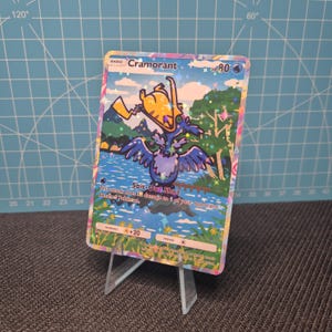 May include: A holographic Pokémon trading card featuring the character Cramorant. The card displays a colorful illustration of the Pokémon with the text "Spit-Out Shot" and other game-related details. The card is propped up on a clear plastic stand.