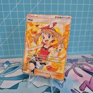 Peut inclure: Une carte Dresseur May holographique du jeu de cartes à collectionner Pokémon. La carte présente une fille de style anime souriante avec des cheveux bruns et un nœud rouge. La carte contient du texte.