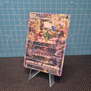 May include: A holographic Pokémon trading card featuring the character Roserade. The card has a colorful floral background and the text "Roserade" at the top. The card is displayed on a clear plastic stand.